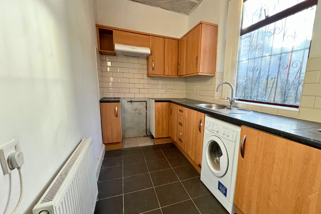 A small kitchen with wooden cabinets, black countertop, a washing machine, a sink with a window above, and tiled backsplash.