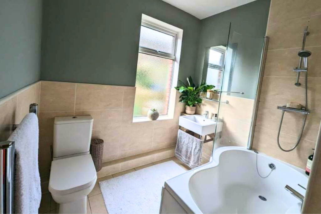 Bathroom with beige tile walls, a white toilet, a bathtub, a sink with a mirror, a window, and a potted plant.