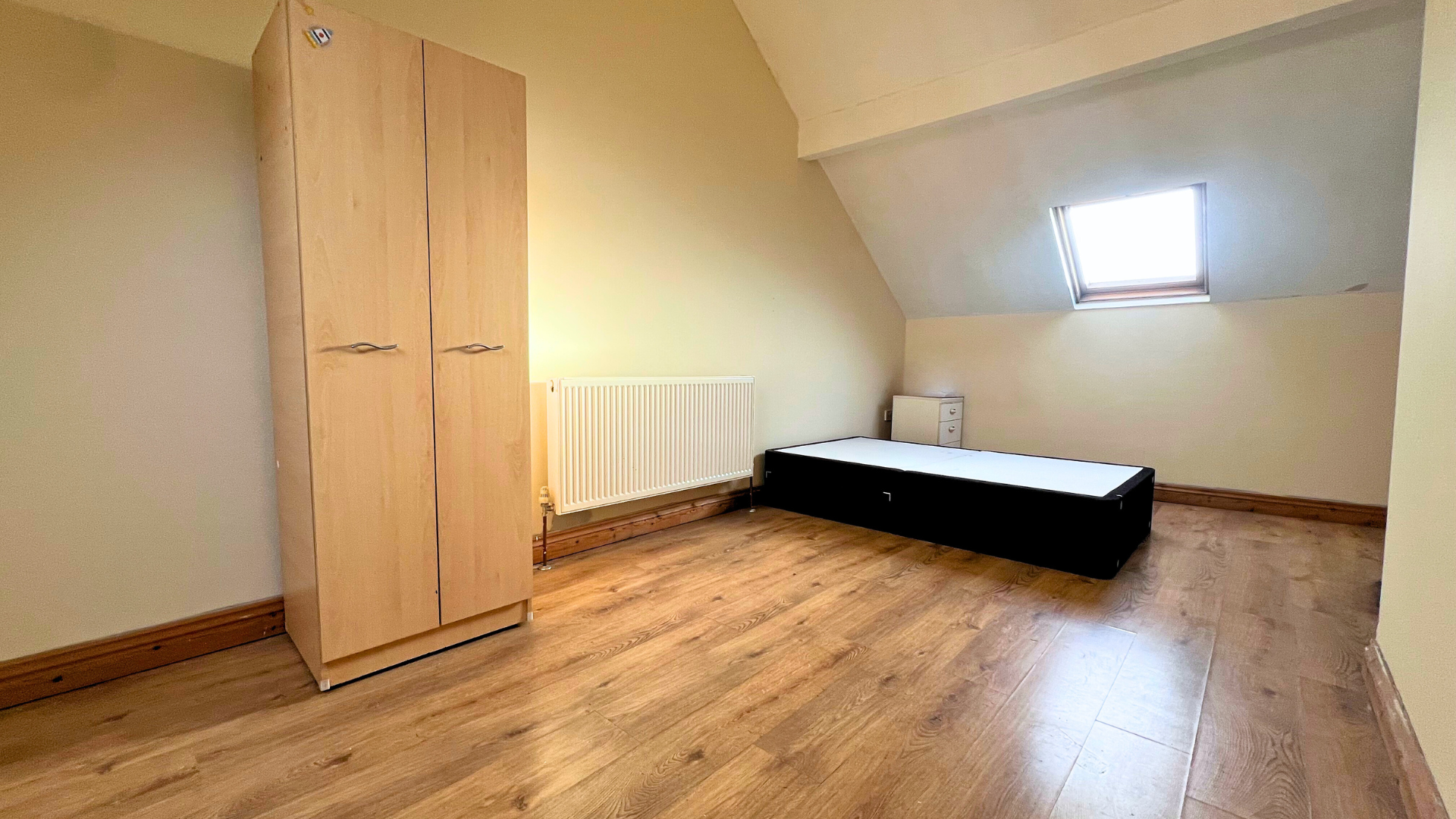 Empty attic room with a wardrobe, a bed frame, a small nightstand, a radiator, and a skylight window.