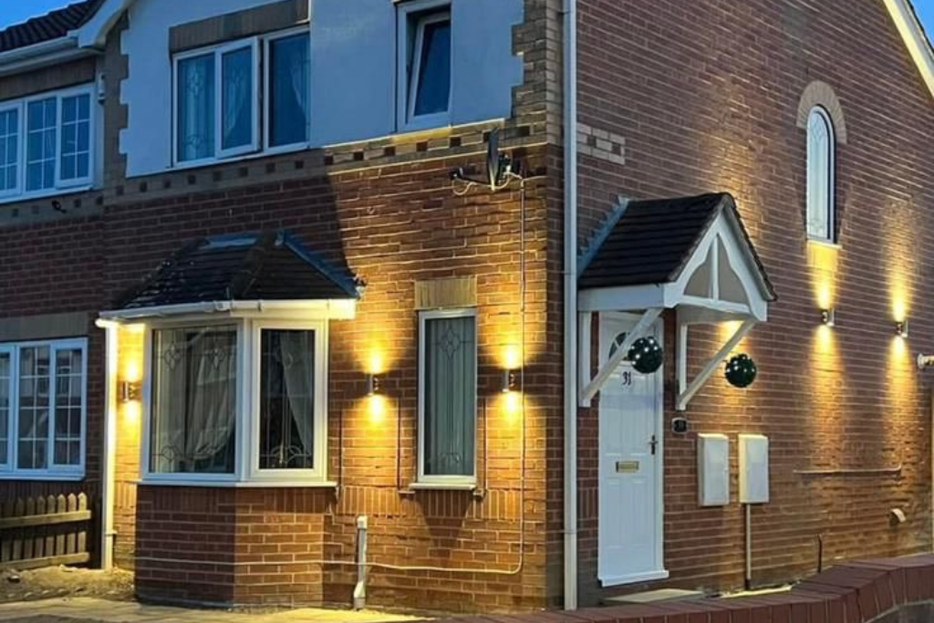 A brick house with exterior lights illuminating the walls at night, featuring a white front door with a small porch, two hanging planters, a bay window with curtains, and a second story with additional windows.