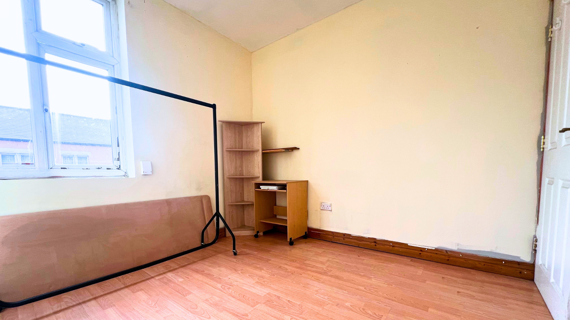 Empty room with wooden floor, beige walls, and a window. Contains a black metal bed frame, a small wooden shelving unit, and an additional small wooden furniture piece. A heating radiator is below the window and a white door is visible on the right.