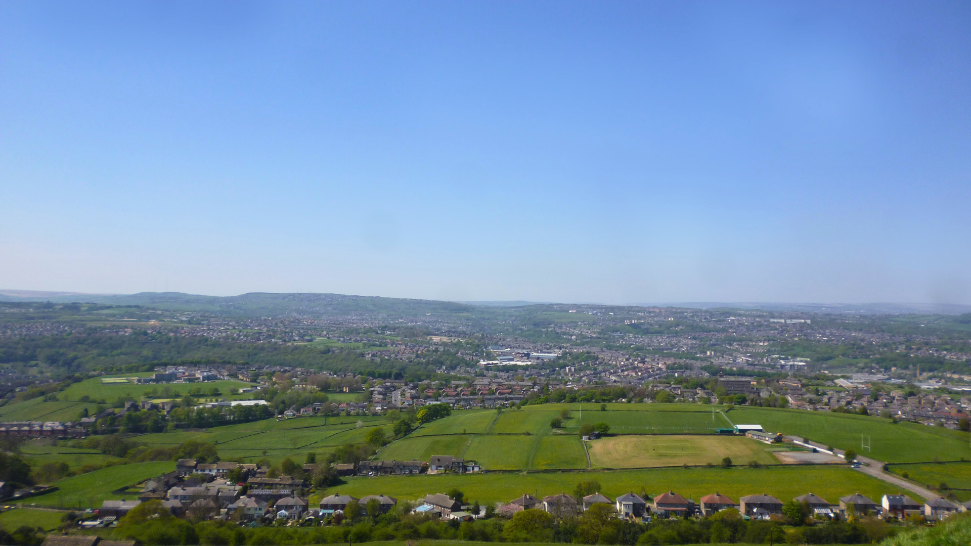 Aerial view of a landscape with green fields, houses, and a distant cityscape under a clear blue sky.