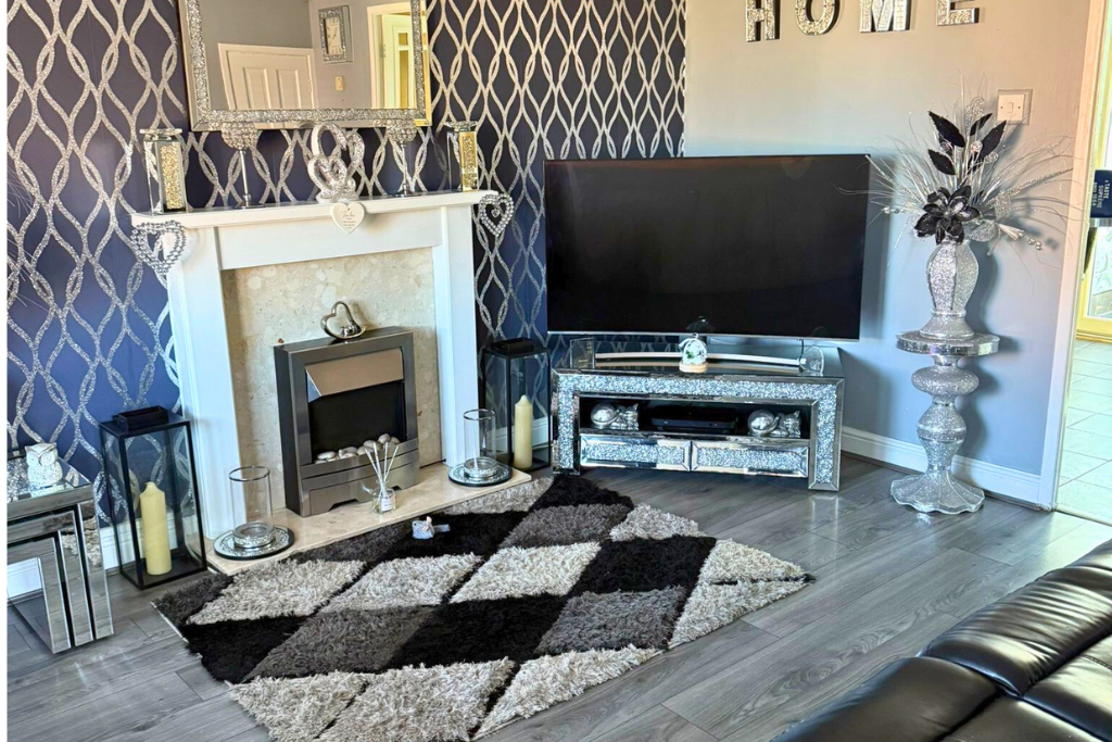 A living room with a fireplace, mirror, TV, and decorative items, including candles and vases, on a dark turquoise wall with a patterned wallpaper background.