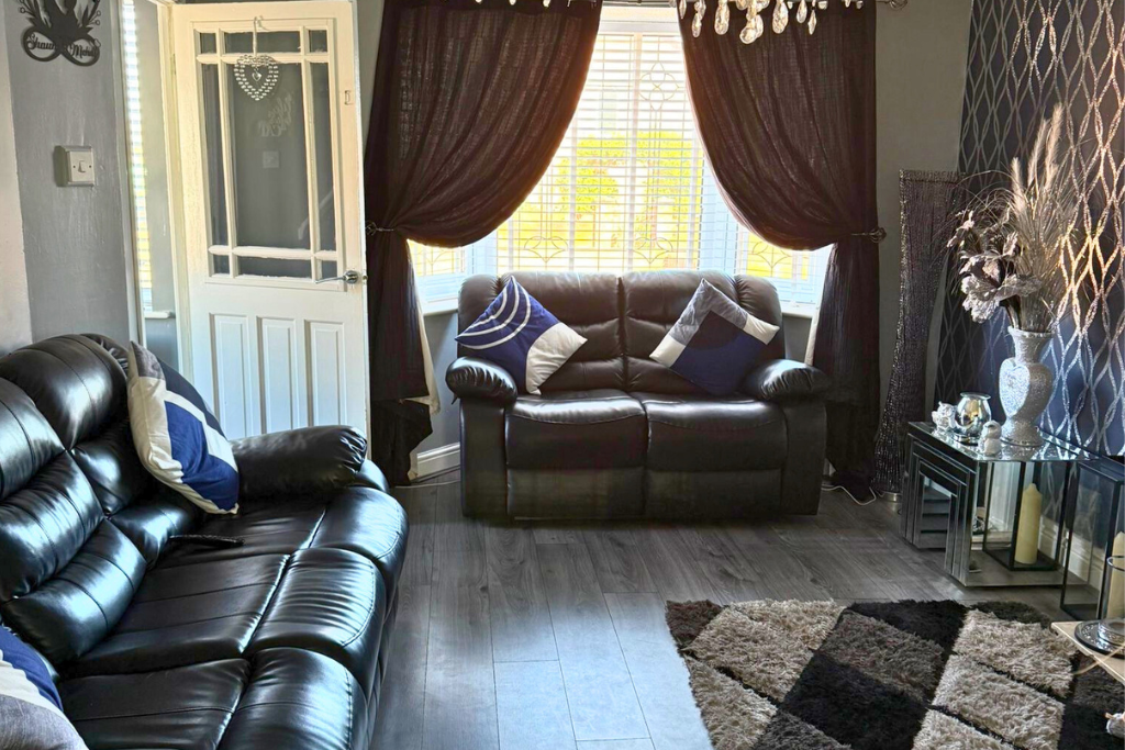 Living room with two black leather sofas, blue and white pillows, large window with white blinds, dark curtains, and a chandelier. There's a glass side table with decorative items and a patterned rug on the floor.
