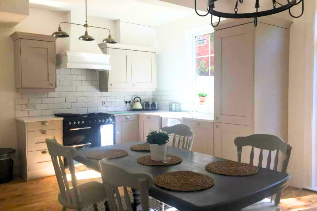Bright kitchen with white cabinets, a black stove, a white tiled backsplash, a round chandelier, a window with a plant, and a dining table with six chairs and woven placemats.