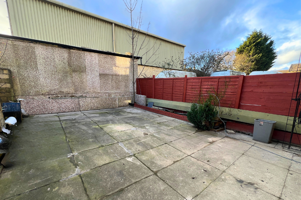 Empty outdoor concrete patio area with a red wooden fence, a small shrub, and neighboring rooftops under a cloudy sky.