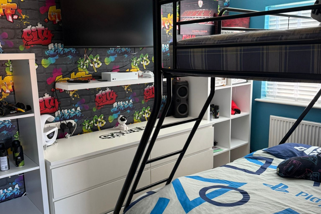 A bedroom with a white dresser, a white shelf, and a blue wall. The bedding has a PlayStation theme, and a bunk bed with a plaid mattress is partially visible. There is graffiti-style wallpaper with colorful cityscape designs on the wall behind the d