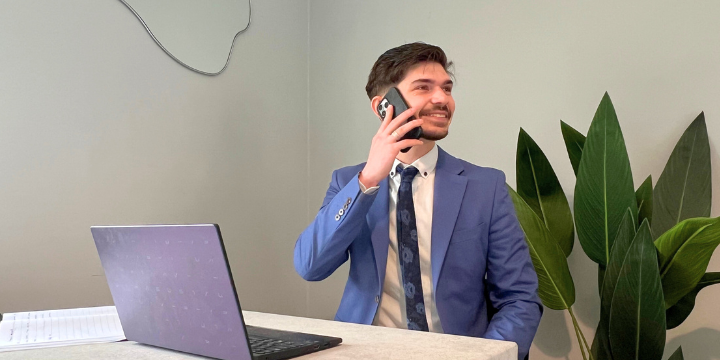 Businessman in a blue suit sitting at a desk with a laptop, talking on a cordless phone, with green plants in the background.