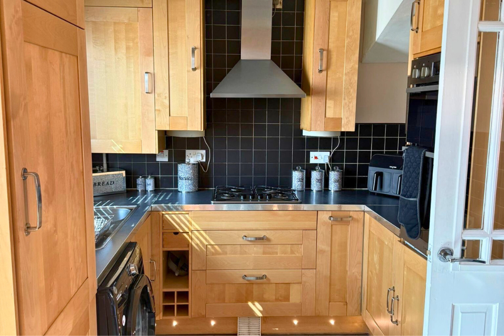 A compact kitchen with wooden cabinets, black countertop, and black tile backsplash. It includes a stainless steel range hood, gas stove, toaster oven, washing machine, and a small wine rack built into the cabinetry.