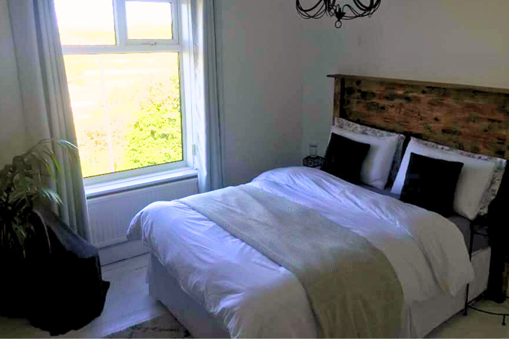 A bedroom with a large window letting in natural light, a neatly made bed with white and dark pillows, a wooden headboard, and a black chandelier hanging from the ceiling.