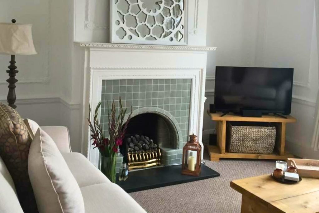 Living room with a white sofa, a fireplace with green tiles, a flat-screen TV on a wooden stand, and a light-colored coffee table.