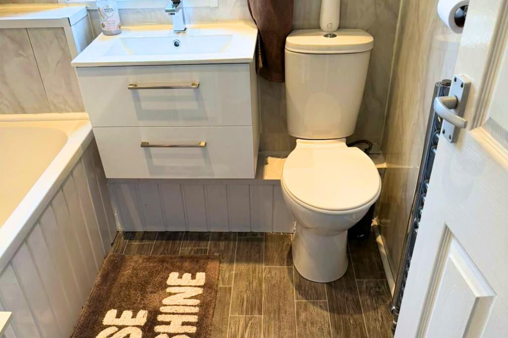 Small bathroom with a toilet, a white sink vanity with two drawers, a brown towel, and a brown rug with the words 'SAVE THE HUMID' partially visible. There is a bathtub on the left and a toilet paper roll on the right.