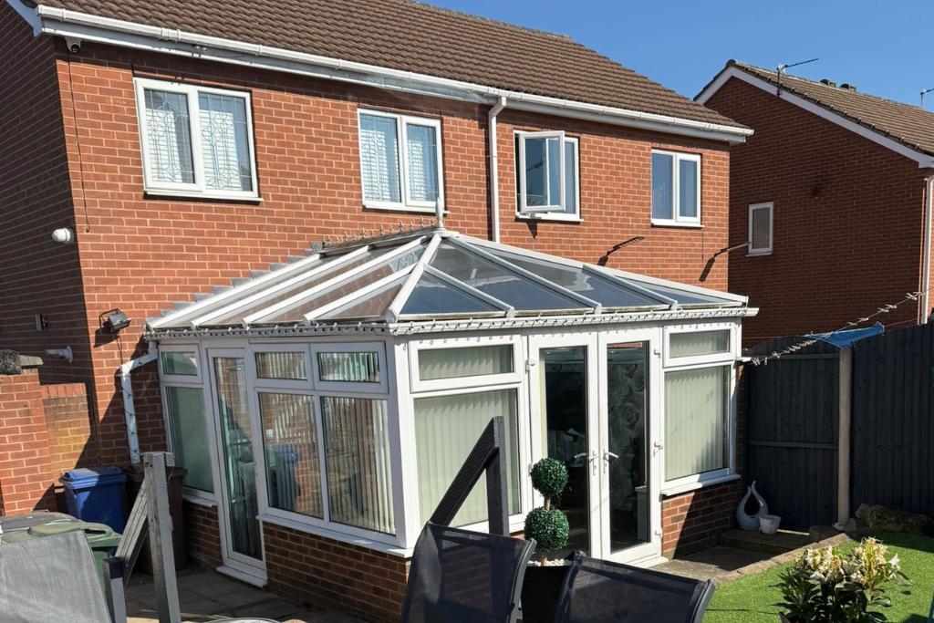 Backyard of a brick house with a conservatory, white-framed glass doors and windows, and a glass roof. There are a few patio chairs, a small bush in a pot, and some decorative items near the conservatory.