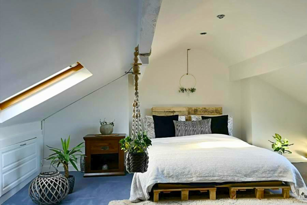 A cozy bedroom with a sloped ceiling, a bed with a wooden pallet frame, black and gray pillows, a hanging plant, a wooden nightstand with a gray vase, and various potted plants near the window
