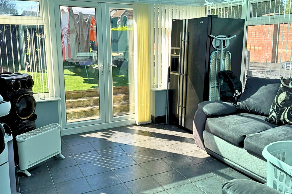 Sunlit room with glass doors leading to a backyard with steps, laundry drying outside, black and silver refrigerator, ironing board, backpack, gray sofa with patterned pillows, black speaker system, heater, and laundry basket.