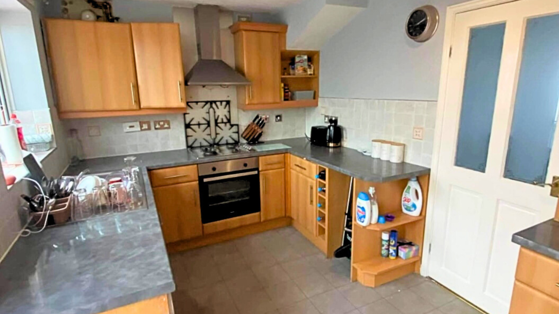 Kitchen with wooden cabinets, gray countertops, a stove, a microwave, and various kitchen items, including dish soap bottles and a dish rack, near a door with frosted glass panels.