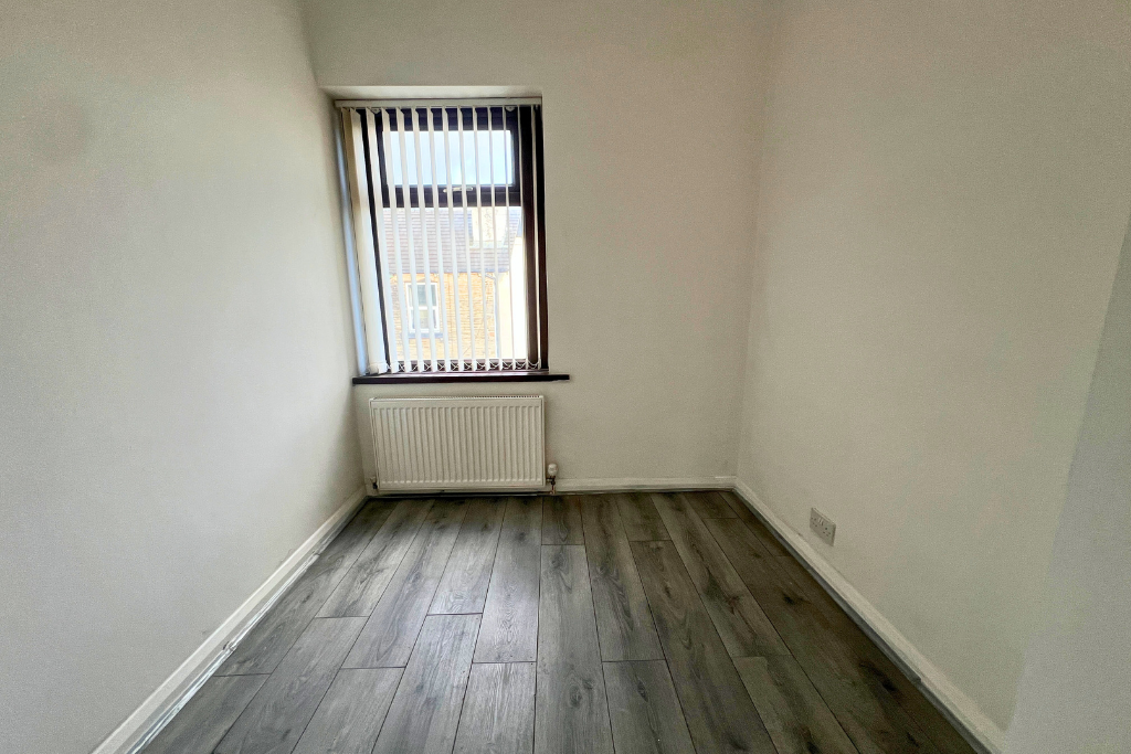 Empty room with white walls, grey wooden floor, a window with vertical blinds, and a radiator below the window.
