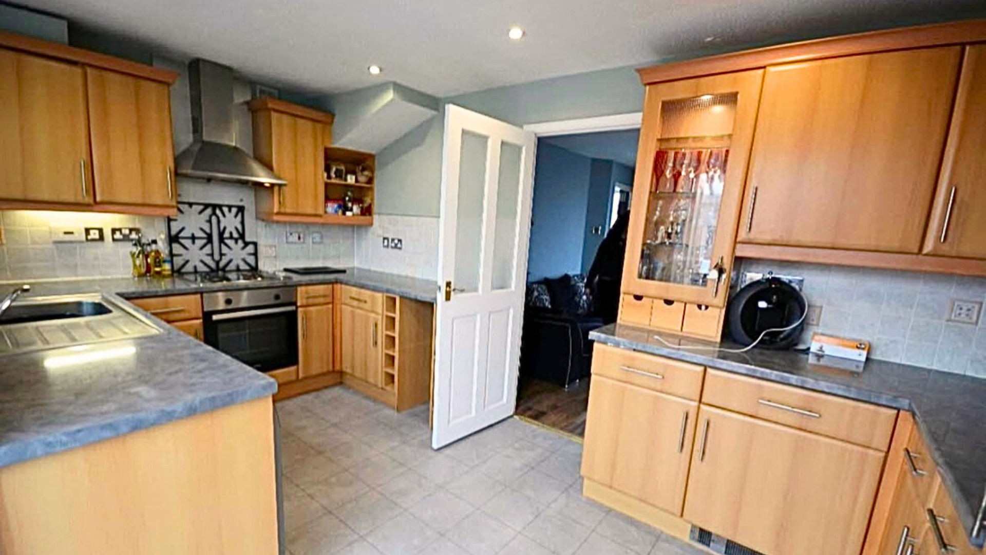 Kitchen with wooden cabinets, gray countertops, tiled backsplash, black stove with stainless steel hood, and open door leading to a living room with black furniture.