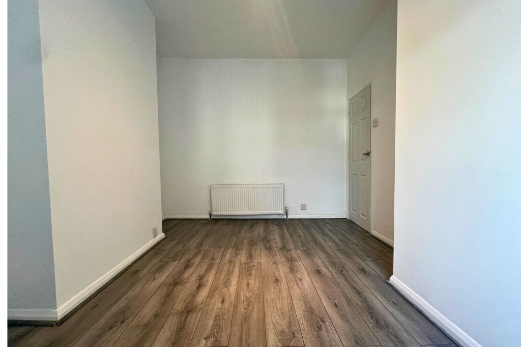 Empty room with wooden flooring, white walls, a white radiator, and a closed white door.