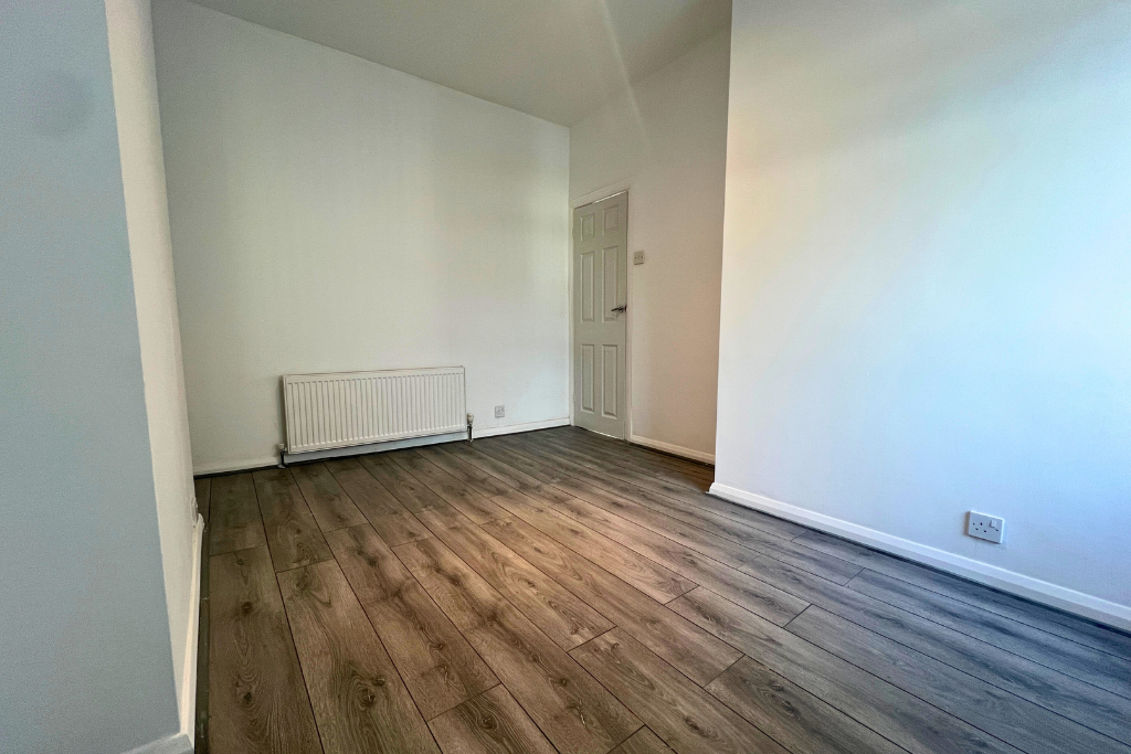 Empty room with white walls, hardwood flooring, a radiator under the window, and a closed door on the far wall.