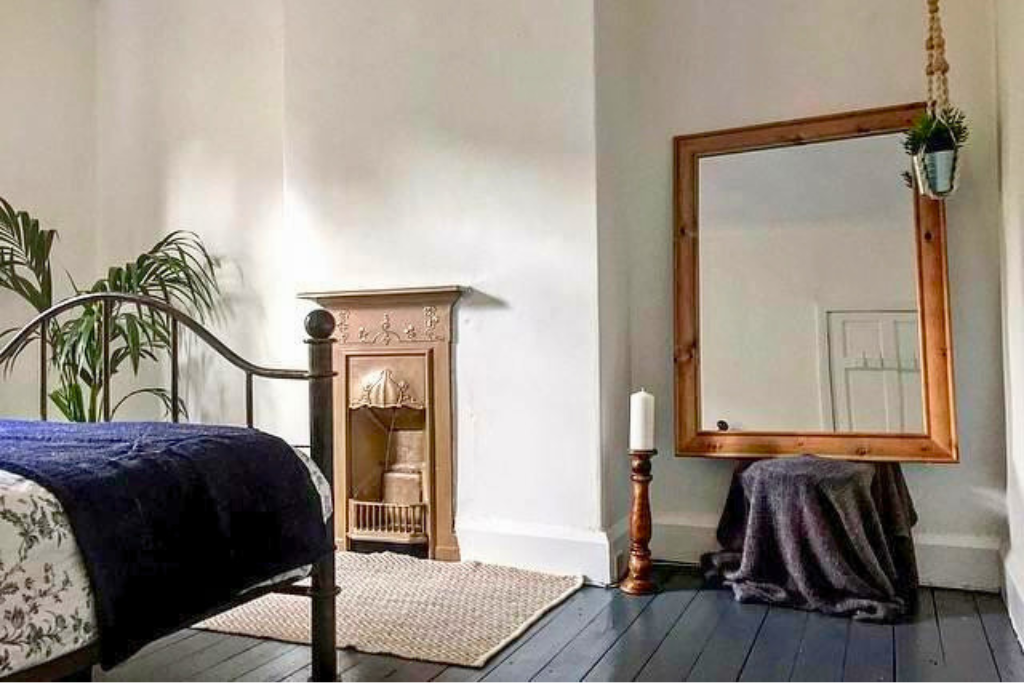 A bedroom corner with a black metal bed, a tall green plant, a small ornate fireplace mantel, a large wooden framed mirror on a stand with a dark cloth covering it, a white candle on a tall wooden candle holder, and a hanging plantspot in the top cor