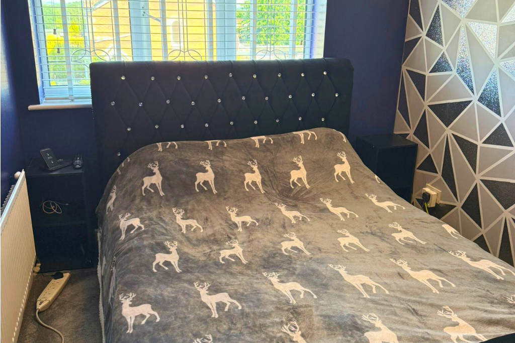 A bedroom with a black diamond-tufted headboard with rhinestone buttons, a window with white blinds, and a duvet cover with white reindeer patterns.