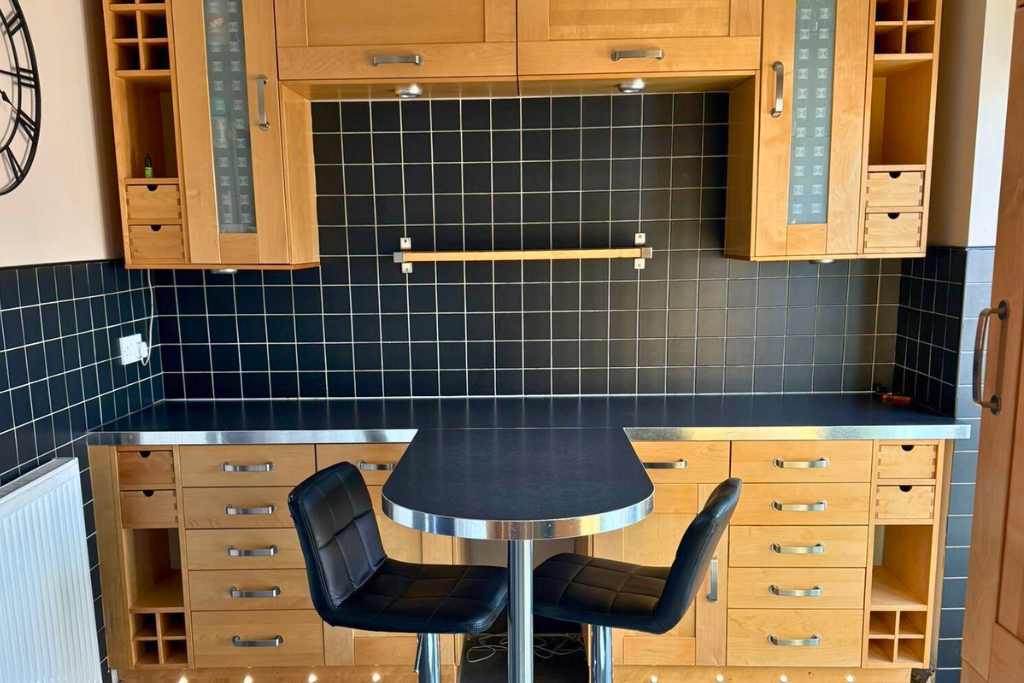 A kitchen with black tiled backsplash, wooden cabinets, and a black countertop. There is a round table with two black chairs in front of the cabinets.