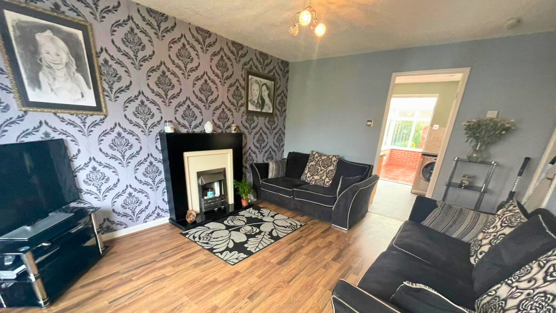 Living room with black and white patterned wallpaper, black sofas with patterned cushions, a black and white patterned rug, a flat screen TV on a black stand, a decorative fireplace with items on top, and a doorway leading to a laundry area with a wa