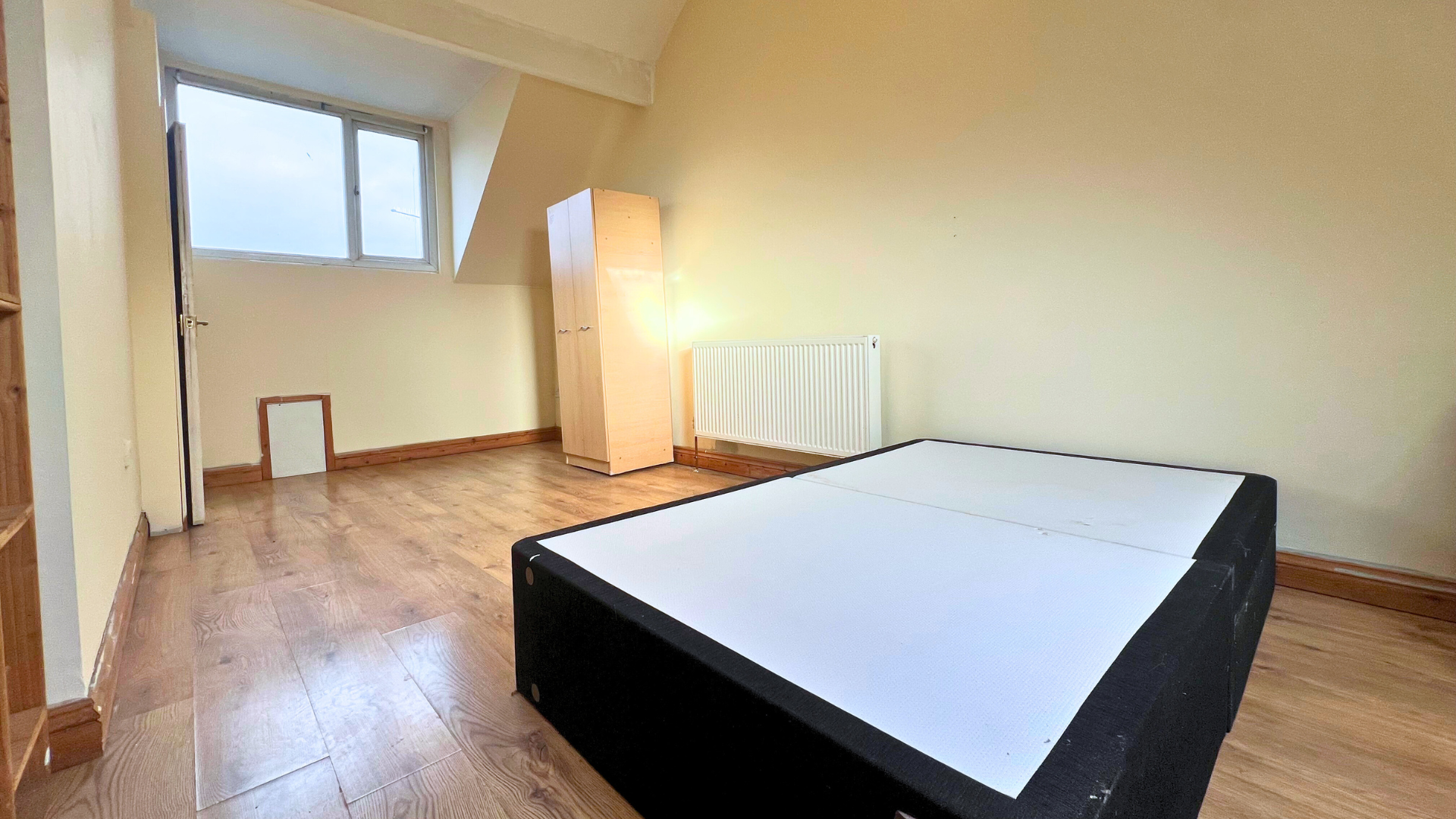 Empty room with hardwood floor, yellow walls, a window, a radiator, a beige wardrobe, and a bed without bedding.
