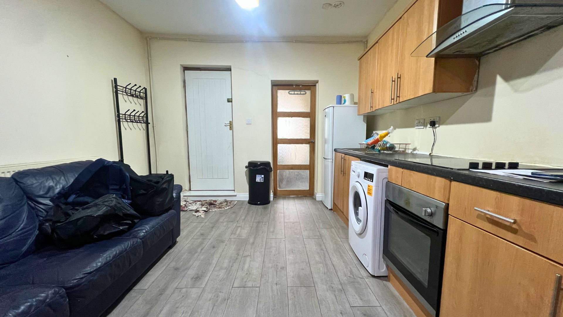 Living room and kitchen with a black leather sofa, backpacks, a door, a door with frosted glass, a fridge, a washing machine, wooden cabinets, a black countertop, a stovetop, and various items on the counter.