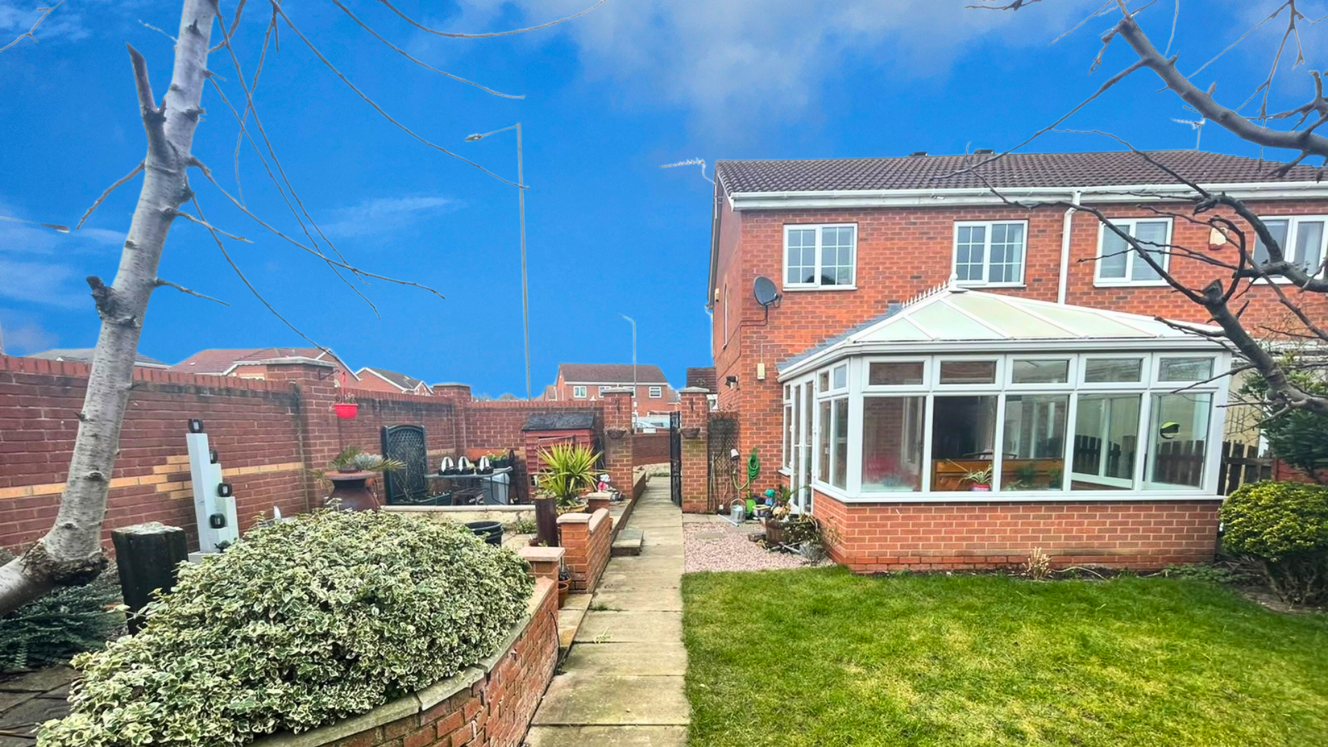 A backyard garden with a concrete pathway, green grass, a brick house with a conservatory, potted plants, outdoor seating, brick walls, and a clear blue sky.