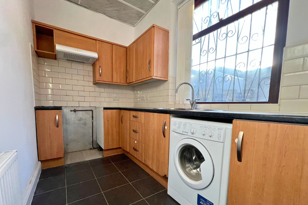 Small kitchen with wood cabinets, black countertop, white subway tile backsplash, washing machine, sink under a window with decorative bars, and a range hood without a stove.