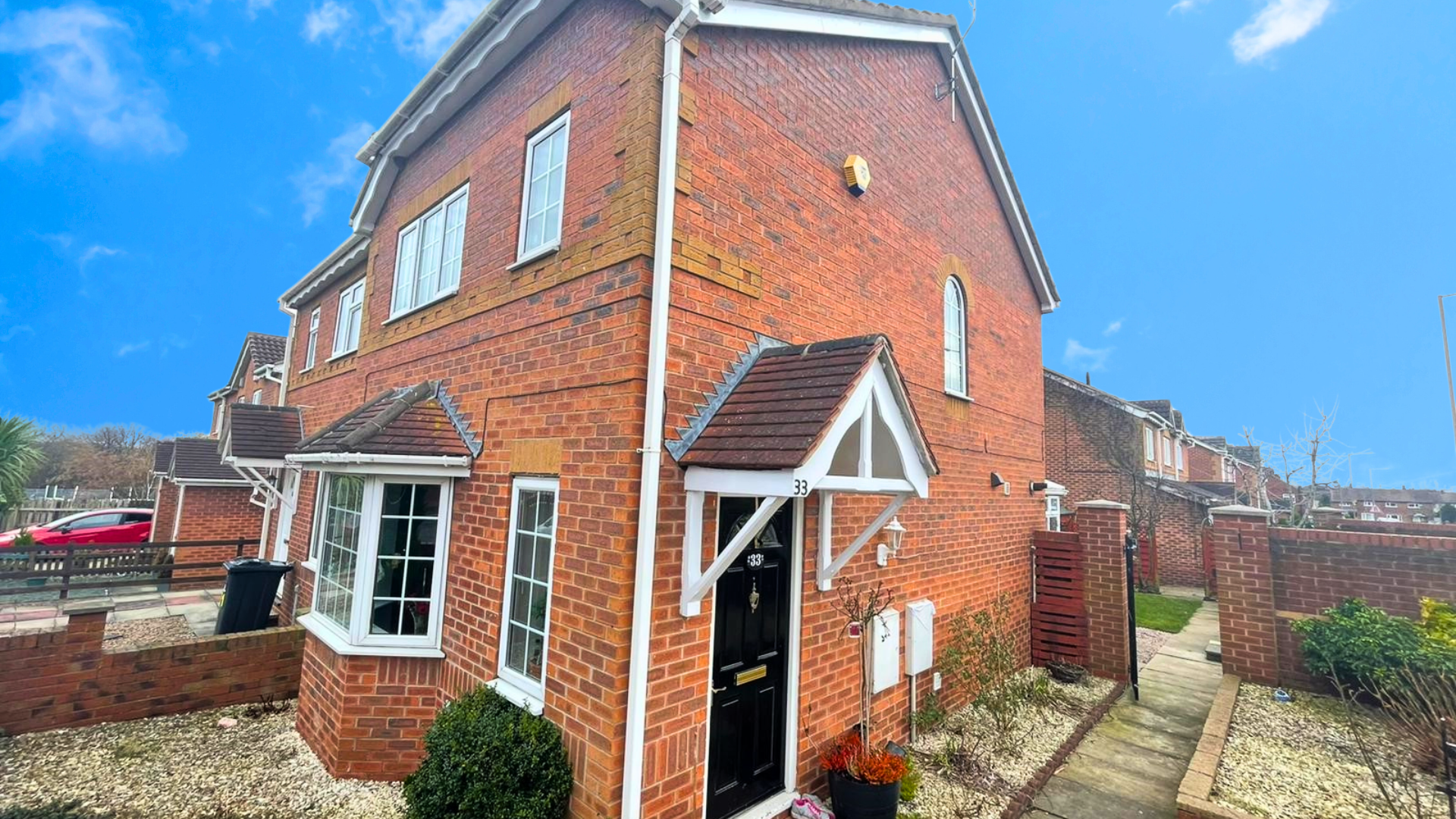 Brick townhouse with black front door, small porch with white trim, bay window, and well-kept garden path, against a bright blue sky.