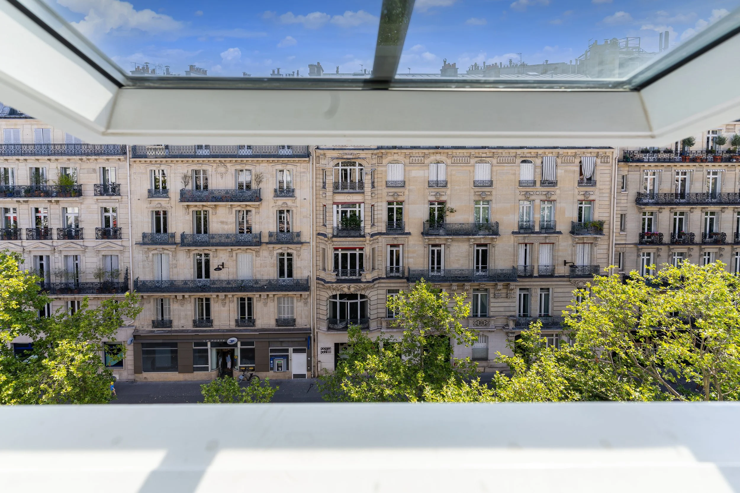 View from a window looking out onto a multi-story Parisian building with balconies and greenery below, under a partly cloudy sky.
