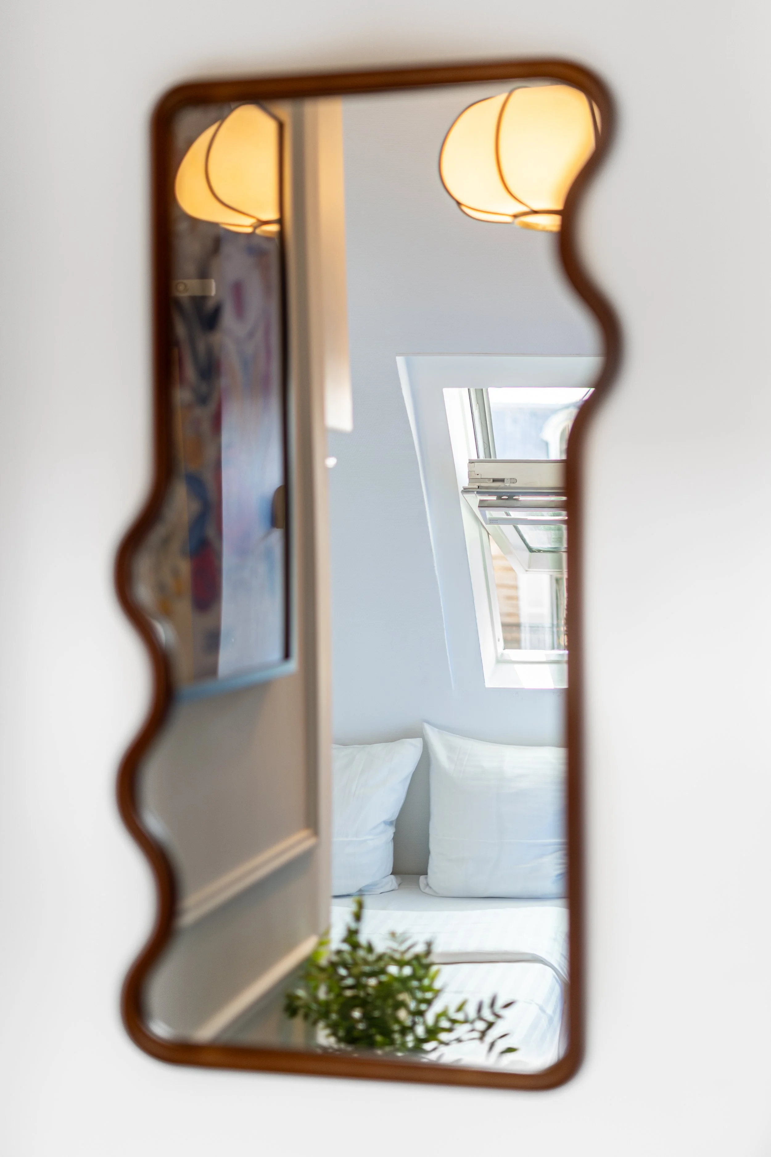 A mirror with a decorative, wavy frame reflecting a bedroom with two pillows, a window, and hanging lamps.