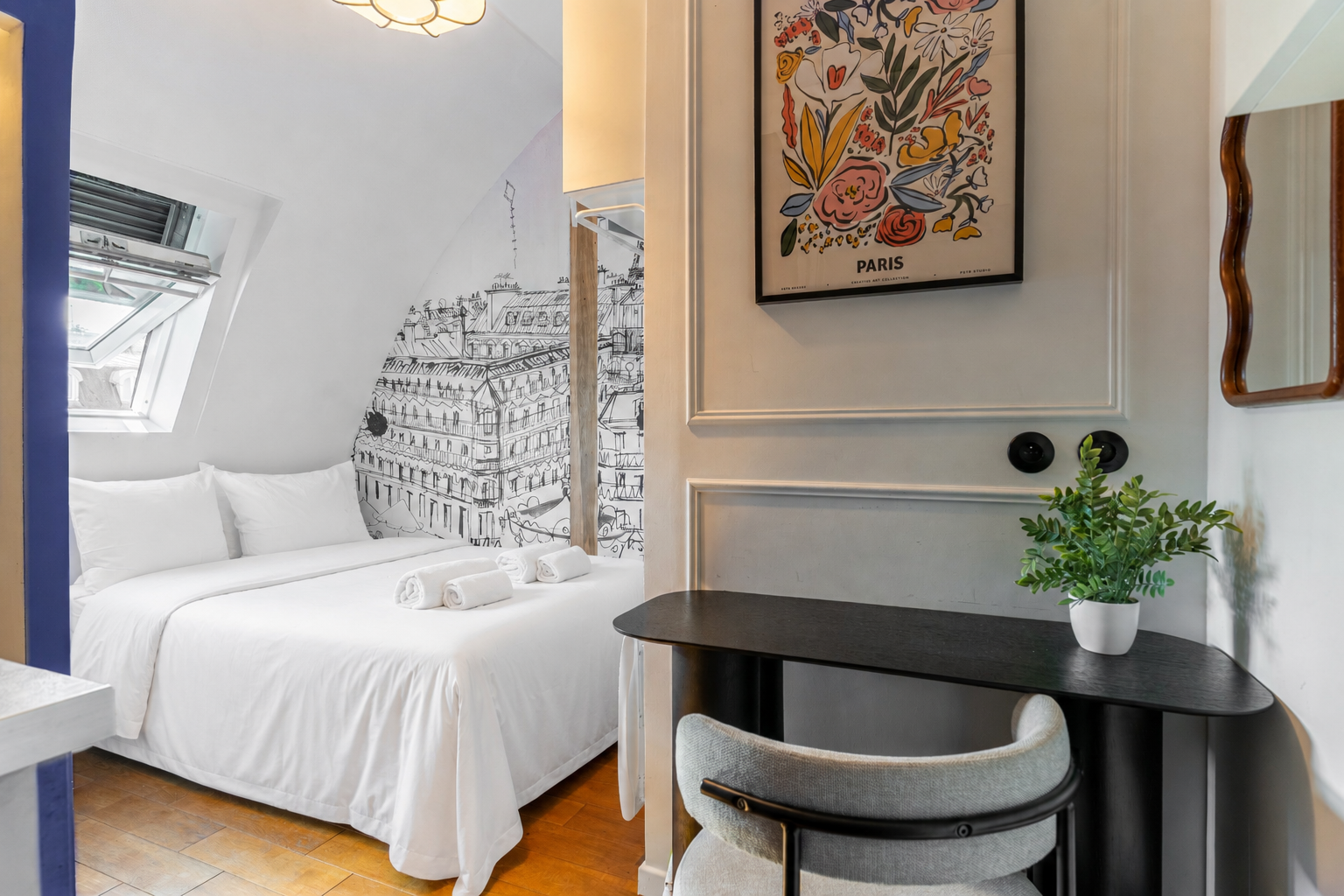 A cozy bedroom with a white bed and linens, decorative black and white art of Paris on the wall, a black table with a green plant, and a window with blinds.