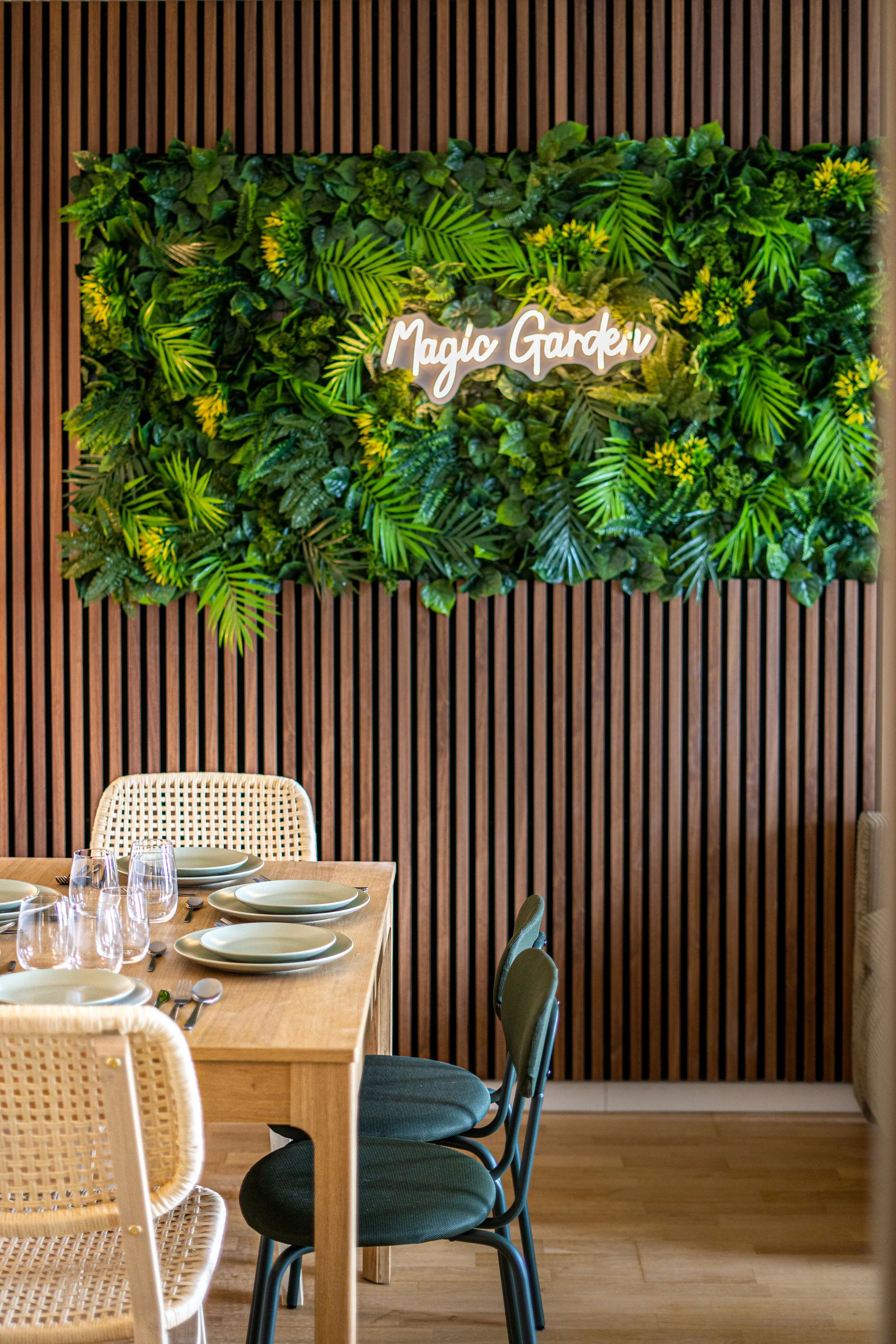 A dining area with a wooden table set with plates, glasses, and silverware, surrounded by chairs with woven or cushioned seats. A wall with a wooden slat design and a green leafy decoration with a sign reading 'Magic Garden' in light-colored cursive lettering is visible in the background.