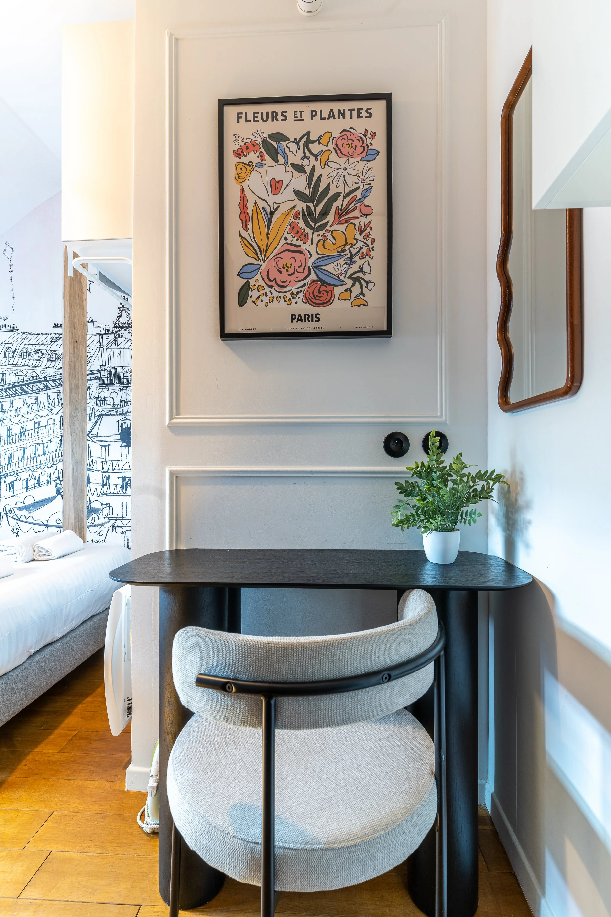 A cozy corner of a room featuring a black round table with a light gray upholstered chair, a potted plant, a wall mirror, and a framed floral poster on the wall. There is also a bed with rolled towels visible to the left, and the room has wooden flooring.