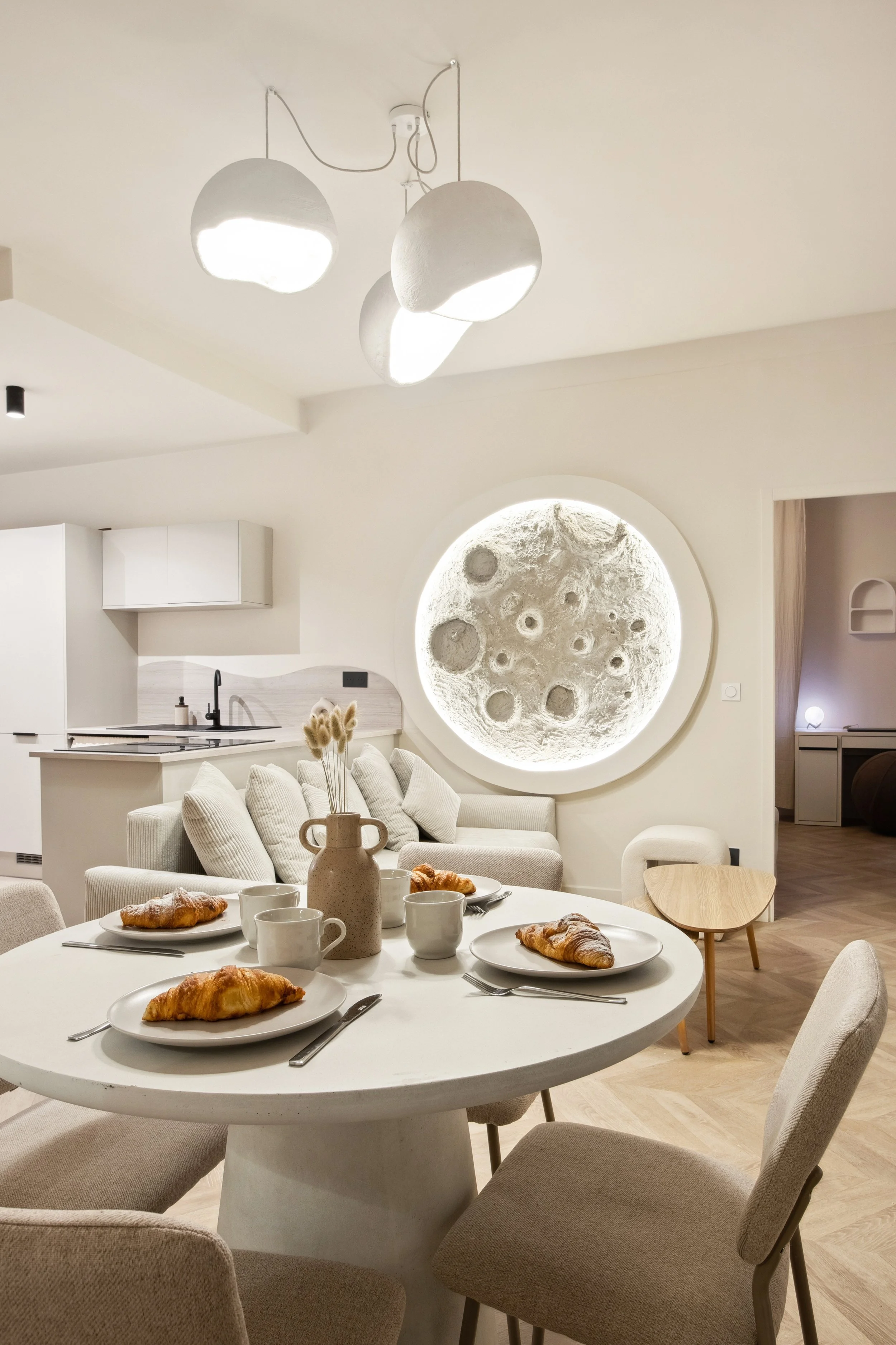 A modern, minimalistic living and dining area with a white round dining table set with croissants, mugs, and a vase with dried flowers. The background features a white kitchen with black fixtures, a large circular moon-like wall decoration with craters, a beige couch with pillows, and a small wooden side table. Overhead, there are contemporary white pendant lights.