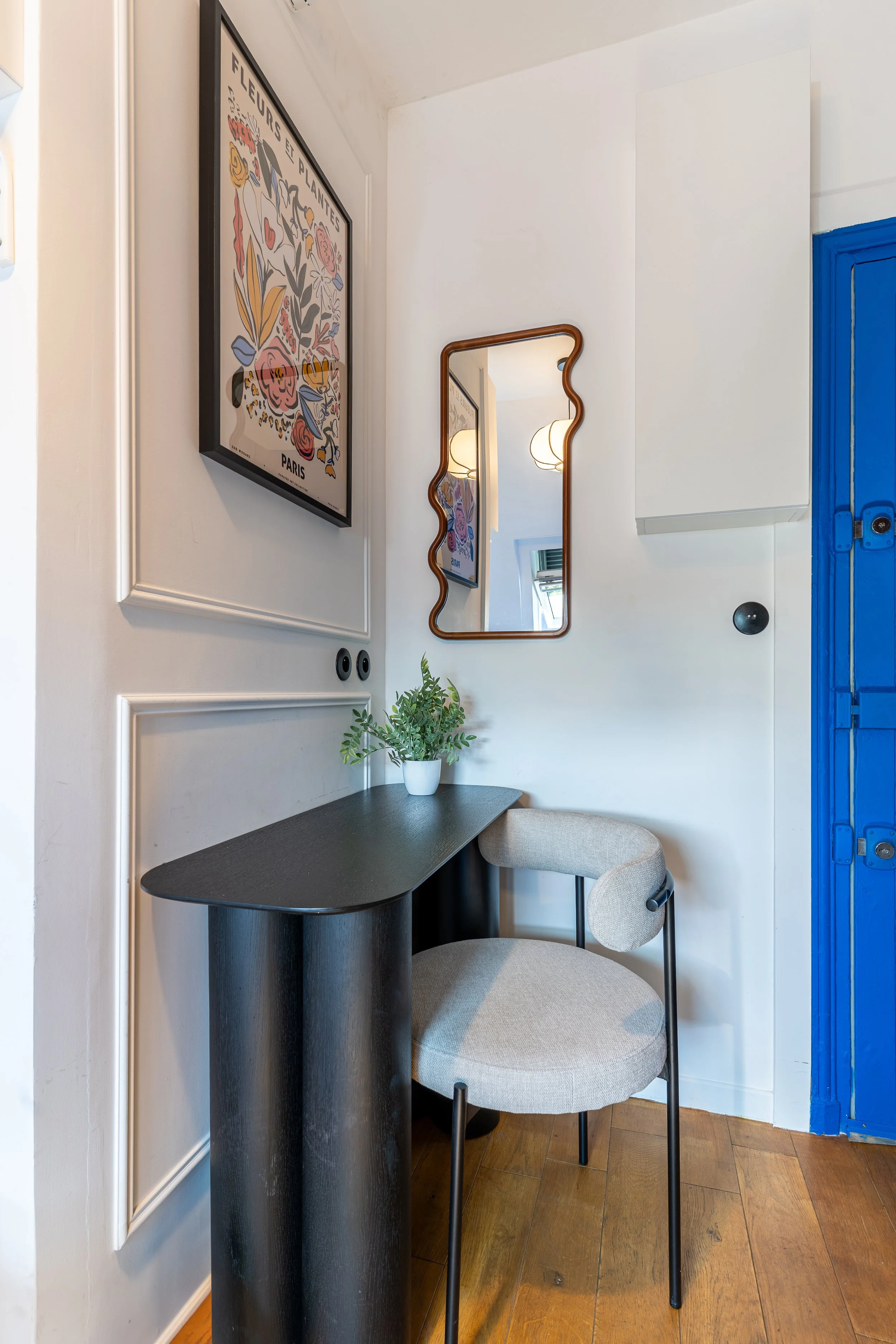 A corner of a room with a black table, a cushioned beige chair, a white wall with decorative molding, two framed art pieces, a small plant, a mirror with a wavy wooden frame, and a blue door.