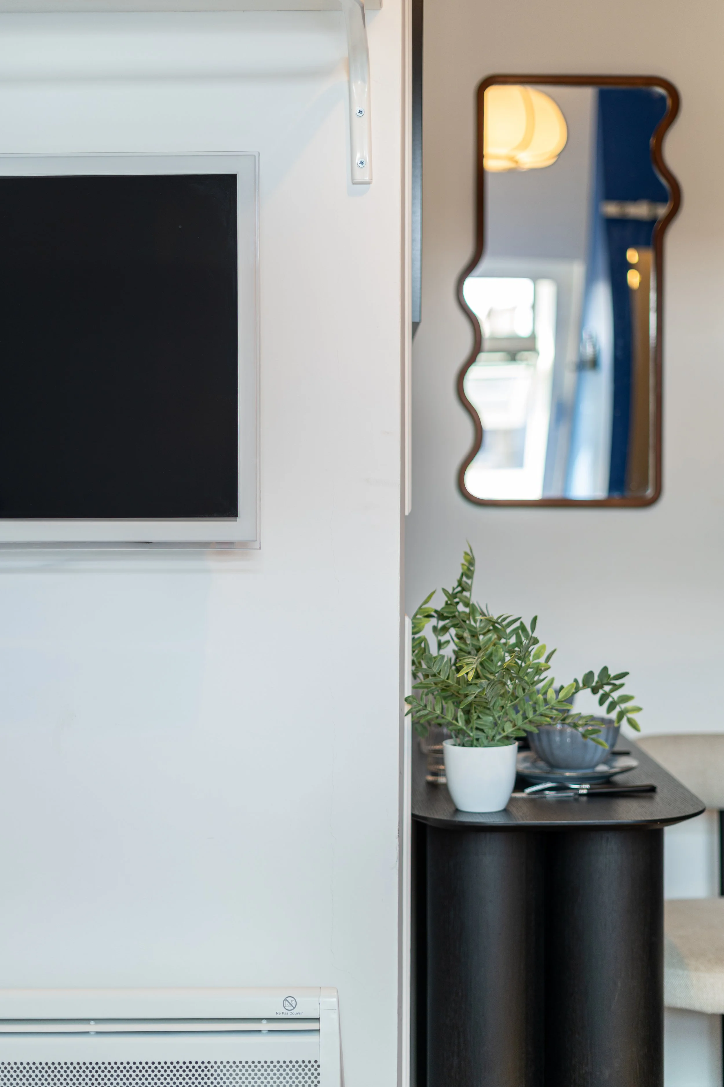 Part of a modern kitchen or living space with a white wall, a wall-mounted blackscreen monitor, and a black table holding potted green plants, a tray, and utensils. A rectangular mirror with a decorative wooden frame is on the wall behind the table.