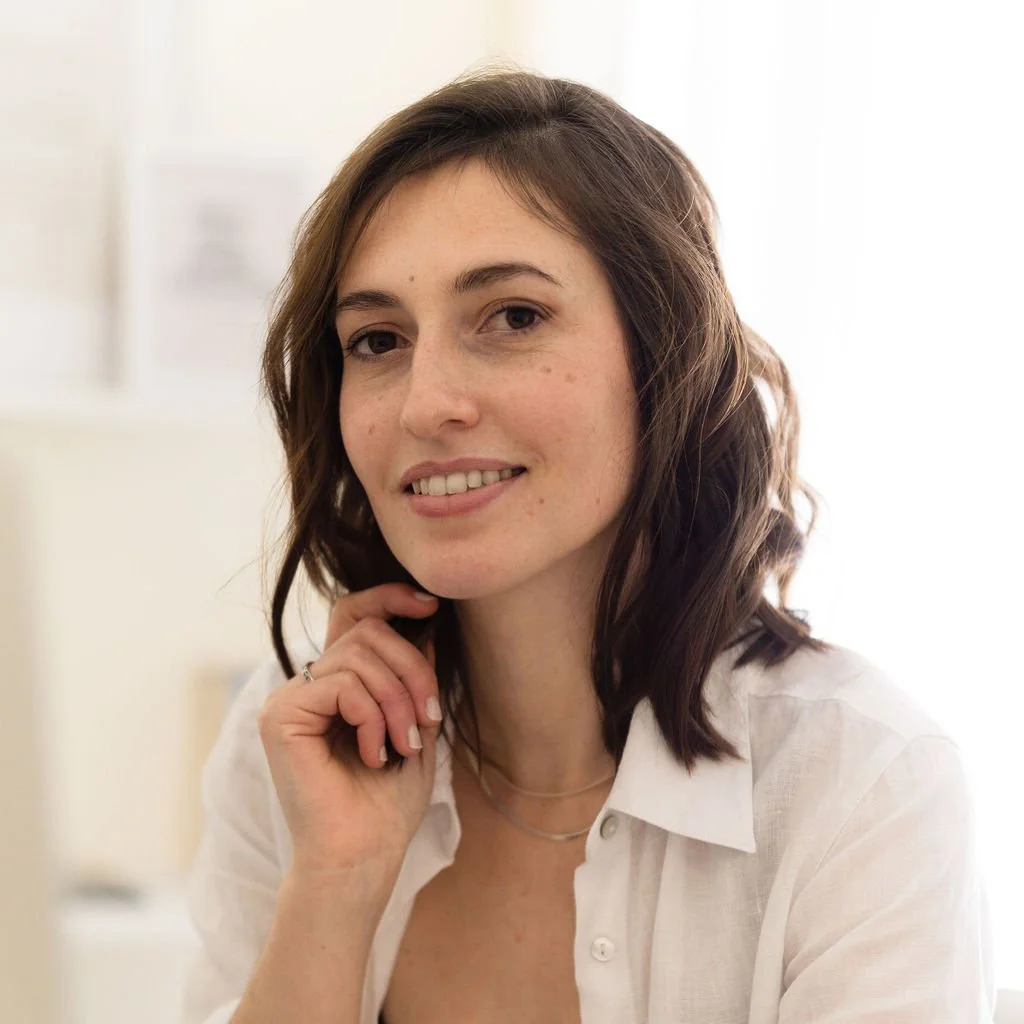 A young woman with shoulder-length wavy brown hair, wearing a white shirt and necklaces, smiling softly with her hand resting near her chin, in a well-lit indoor setting.