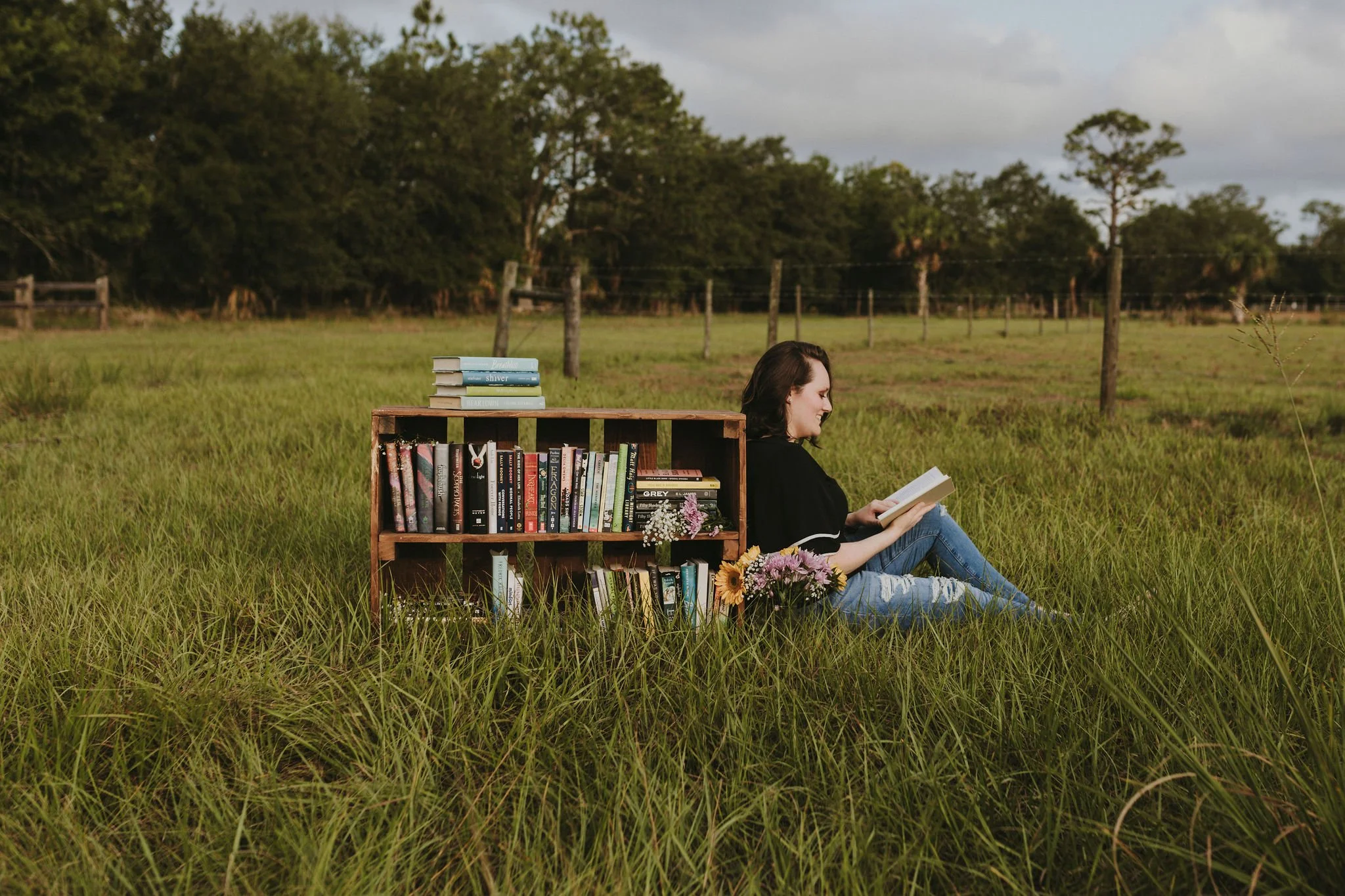 A woman sitting in a grassy field reading a book, a wooden bookshelf filled with books and flowers beside her, trees and fence in the background under a cloudy sky.