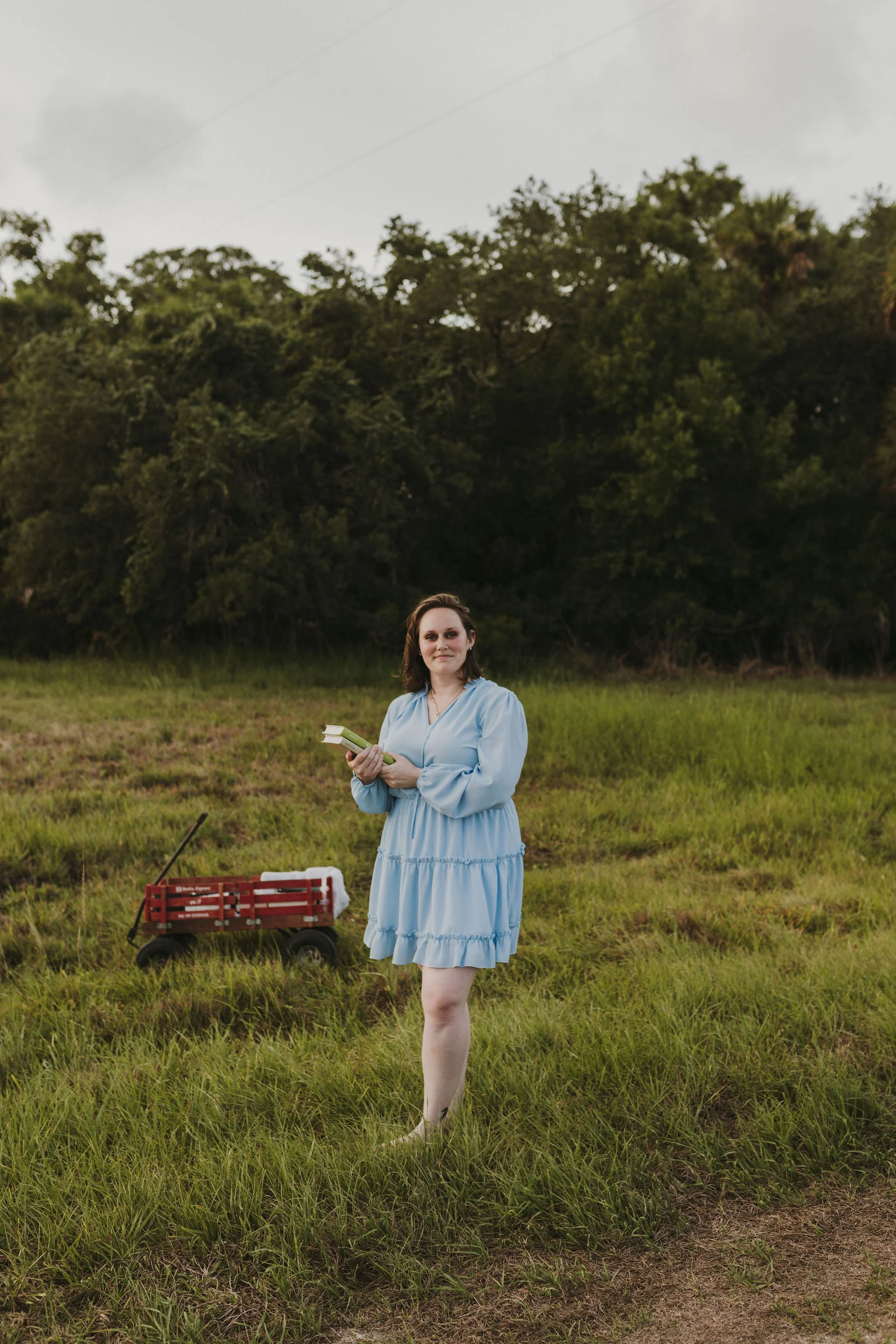 A woman in a light blue dress holding a couple of books standing in a grassy field with trees in the background and a red wagon nearby.