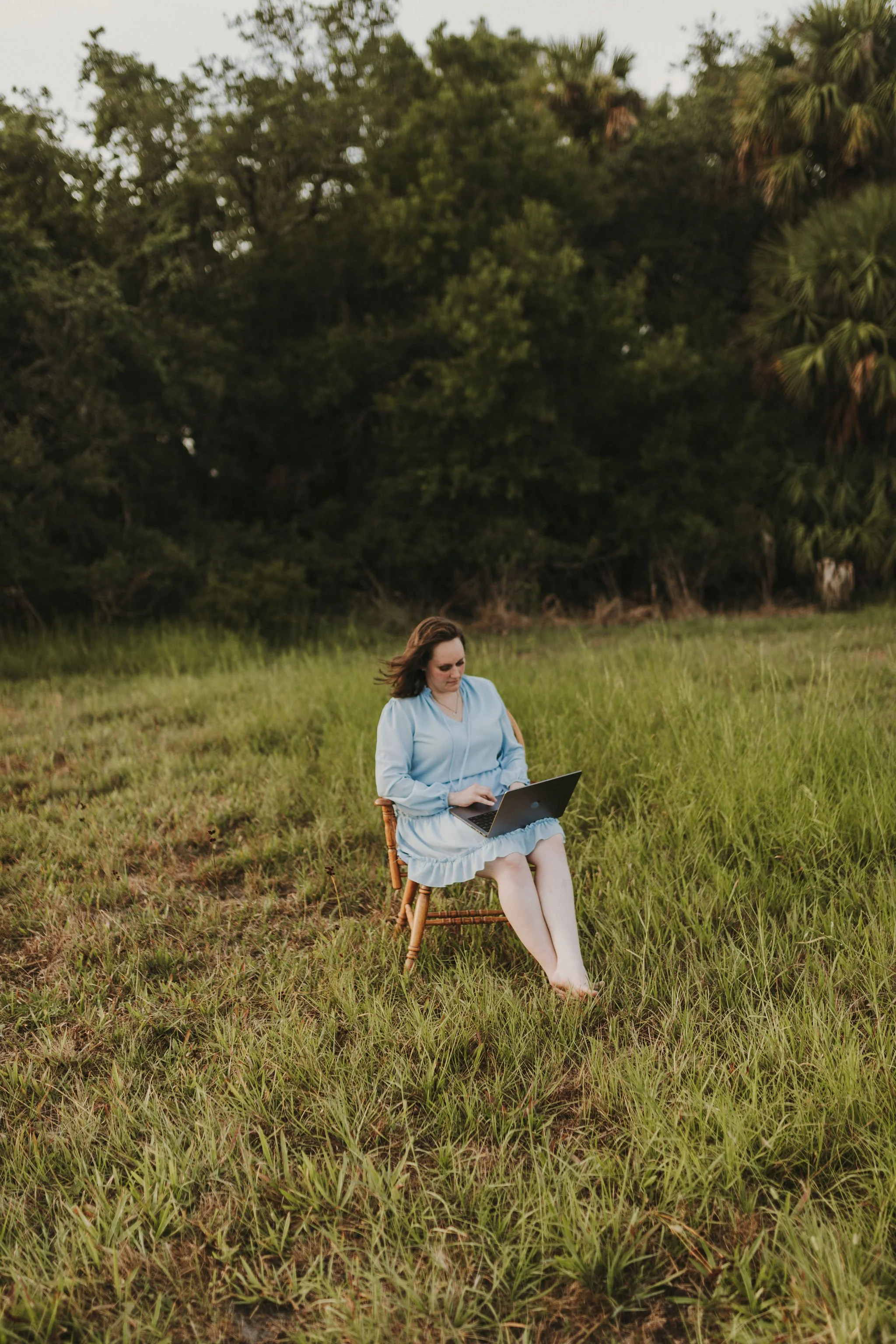 A woman sitting barefoot on a wooden chair in a grassy field, working on a laptop, with trees in the background.