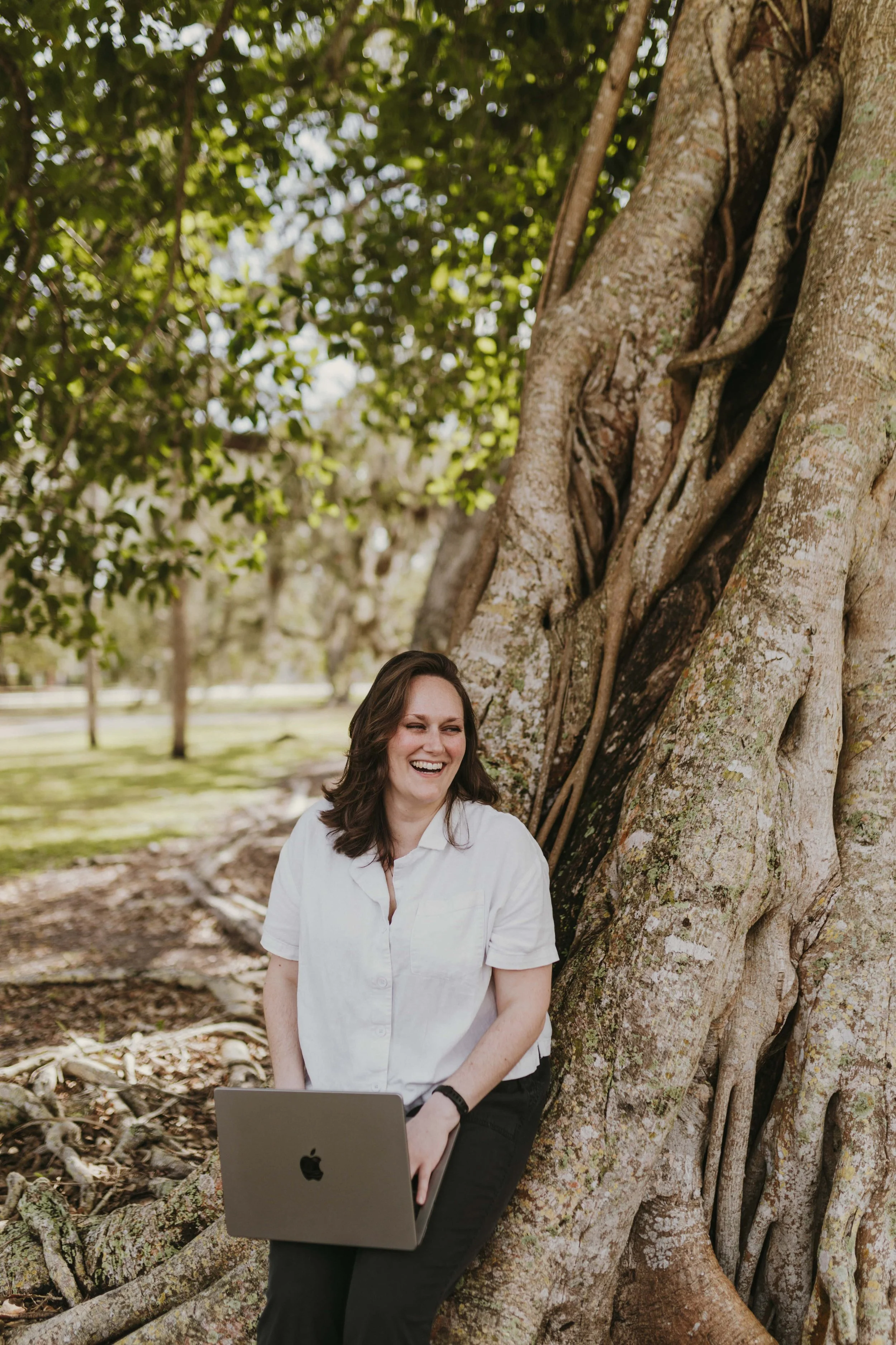 A woman sitting on the roots of a large tree, working on a laptop, outdoors in a park, smiling and laughing.