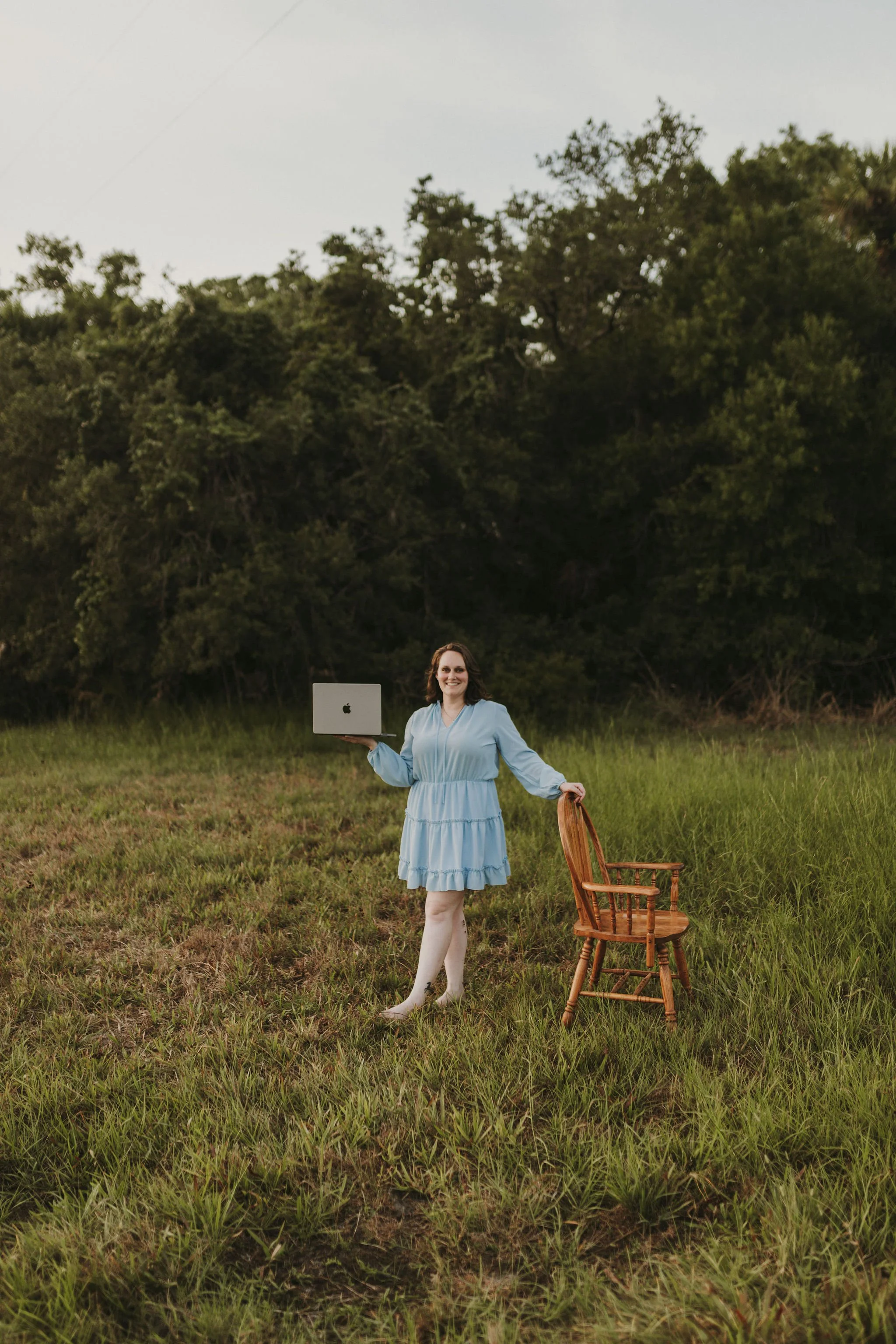 A woman in a light blue dress holding a laptop and a wooden chair standing in a grassy field with trees in the background.