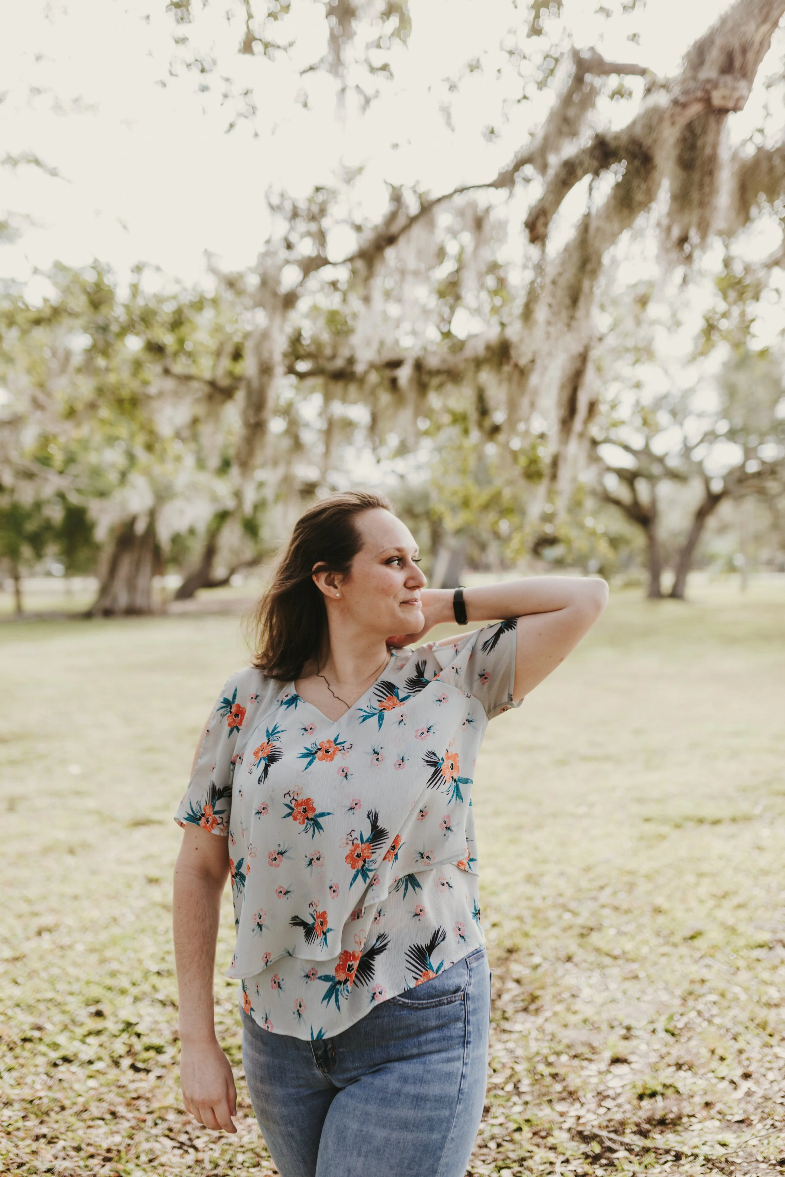 A woman with shoulder-length hair wearing a floral top and jeans standing outdoors in a park with trees and moss hanging from branches.