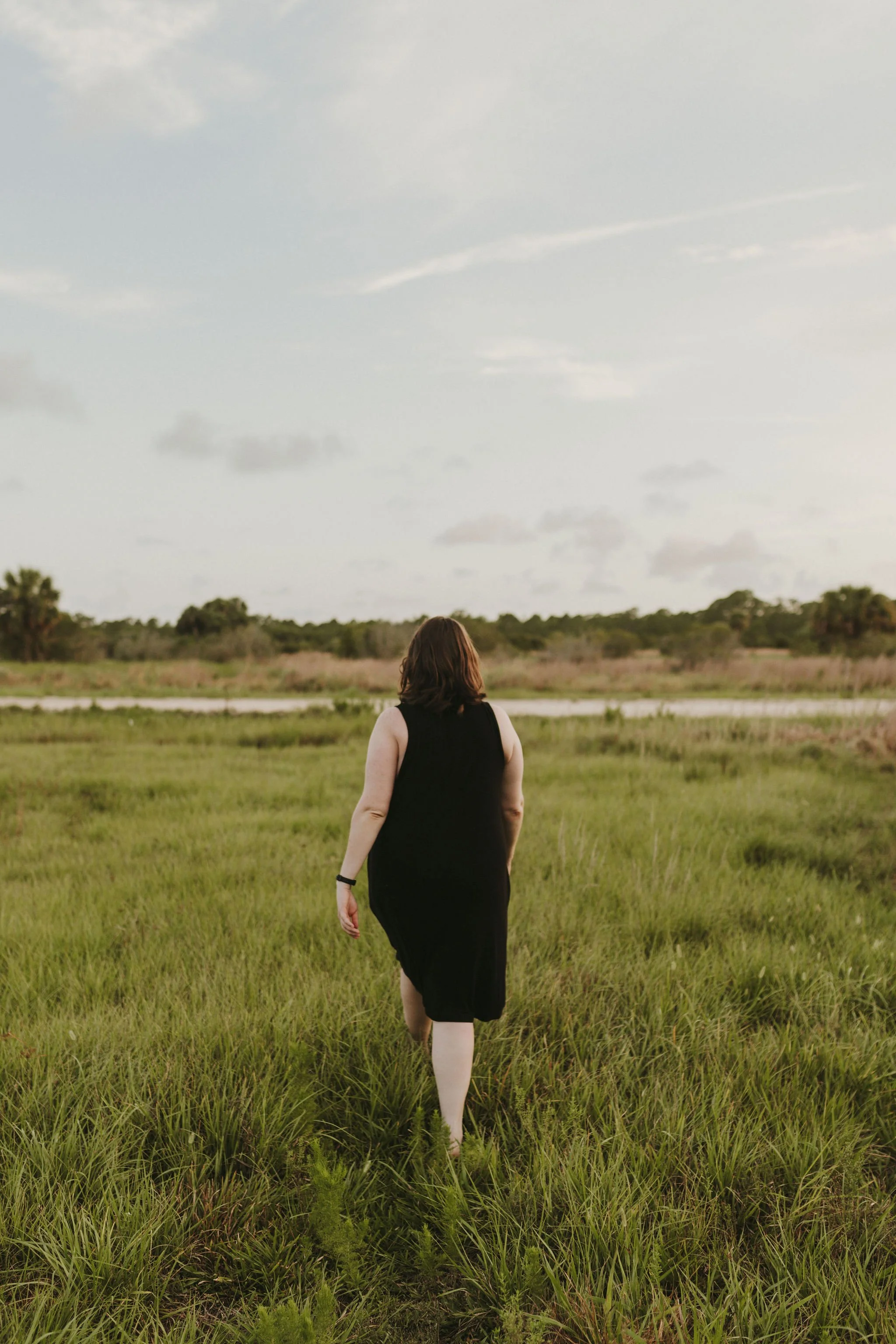A woman in a black dress walking through a grassy field with a cloudy sky overhead.