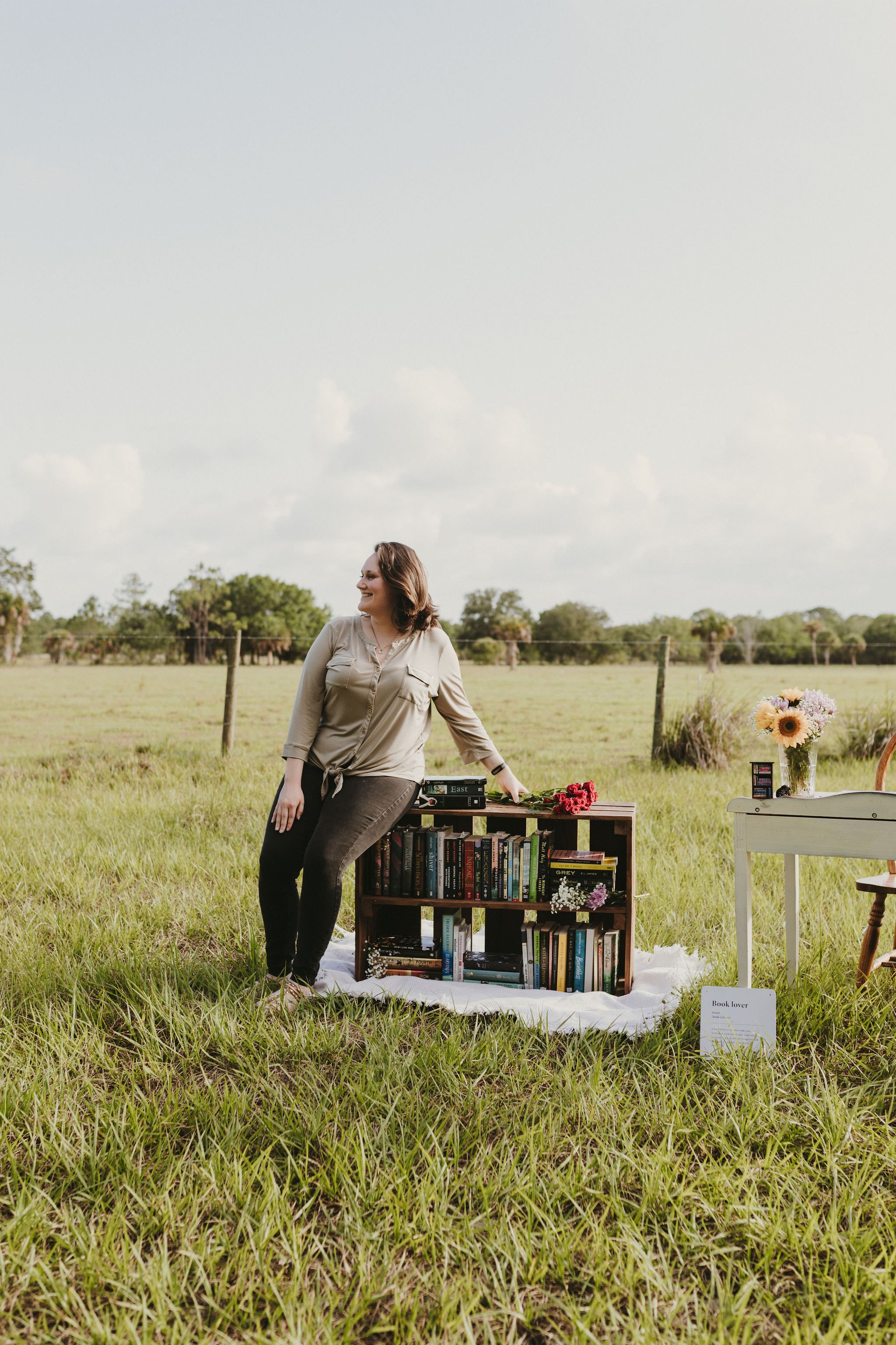 A woman in beige blouse and dark jeans poses outdoors next to a bookshelf on a white cloth, with flowers and a chair nearby, in a lush green field under a partly cloudy sky.